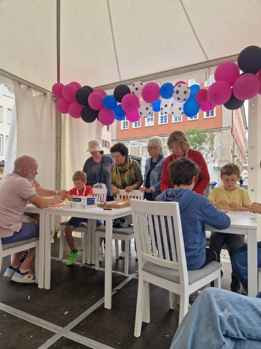 Familien basteln gemeinsam am Tisch im Zelt, darüber eine Ballongirlande in Pink, Schwarz und Blau